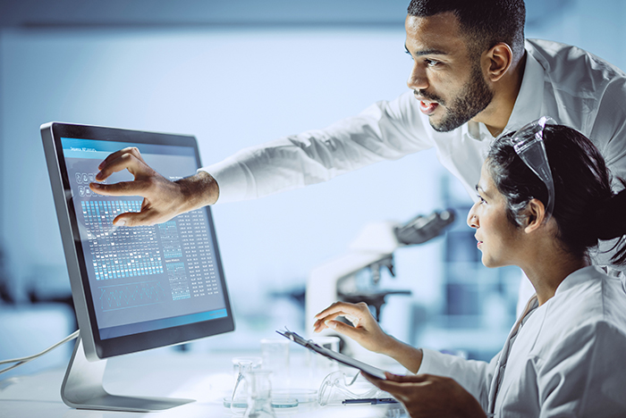 Dos investigadores con batas blancas de laboratorio, uno sentado y otro de pie, miran datos genéticos en un monitor de computadora.